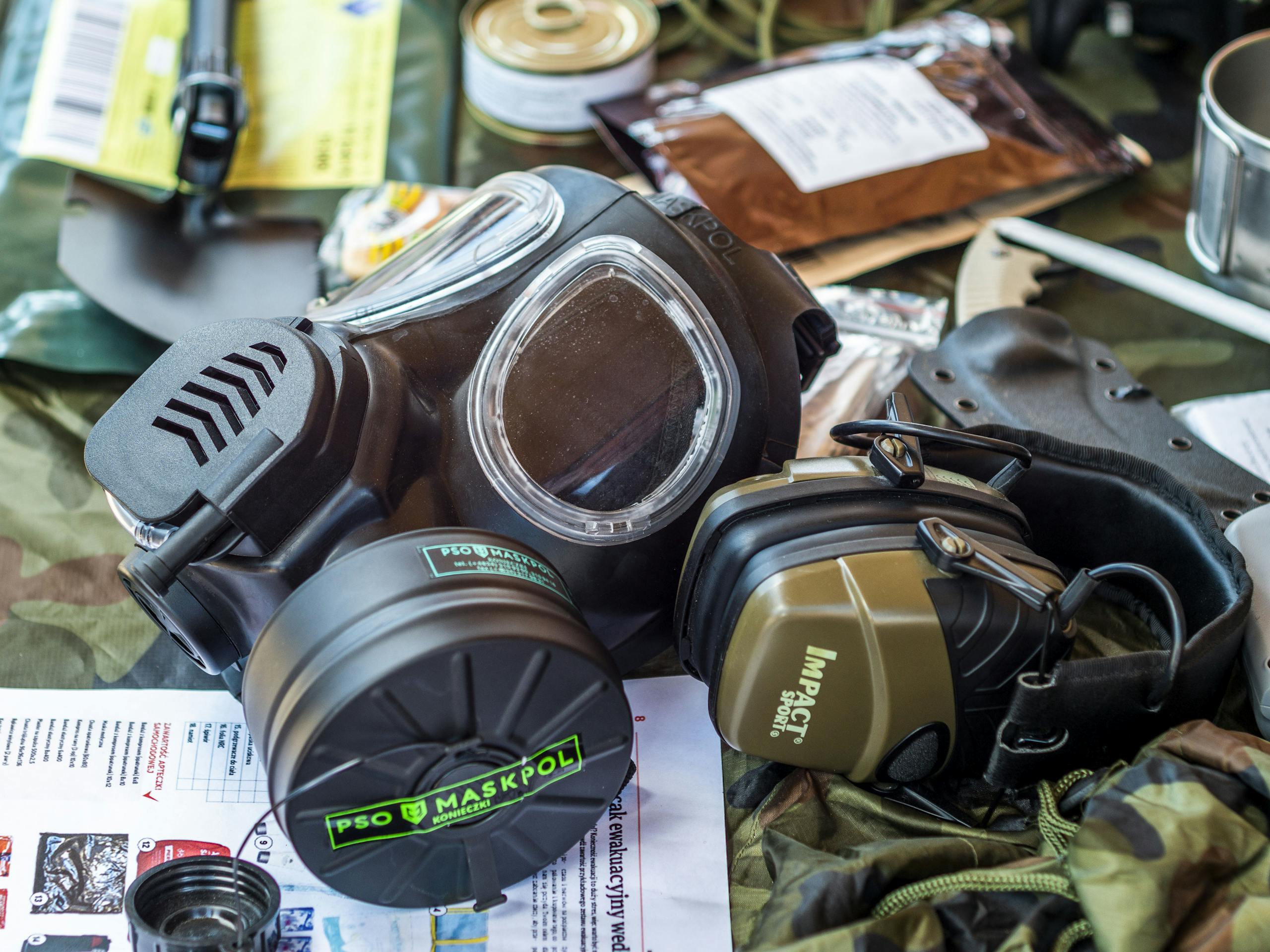 Survival gear including a gas mask and tactical headset on a camouflage background.